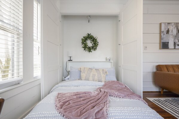 A bedroom with a bed, a window, a wall-mounted wreath, a bedside table, and a chair.