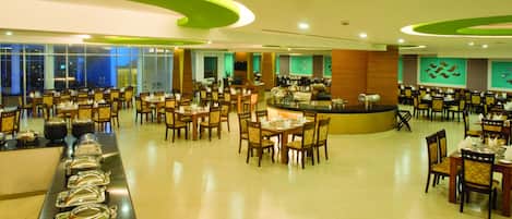 Un comedor amplio con varias mesas y sillas, una barra de buffet con recipientes de comida, y una barra con máquina de café.