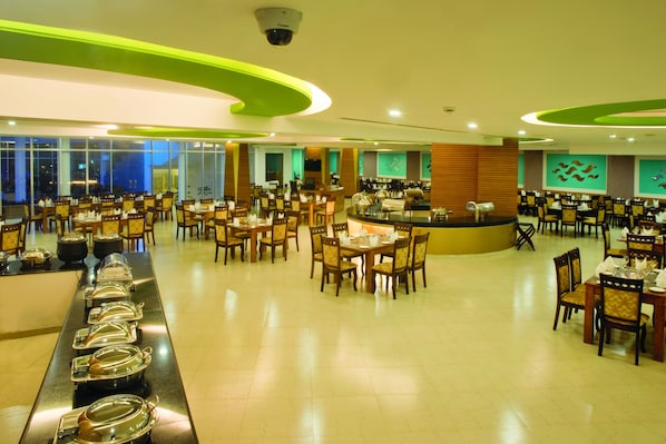 A spacious dining area with multiple tables and chairs, a buffet line with food containers, and a counter with a coffee machine.