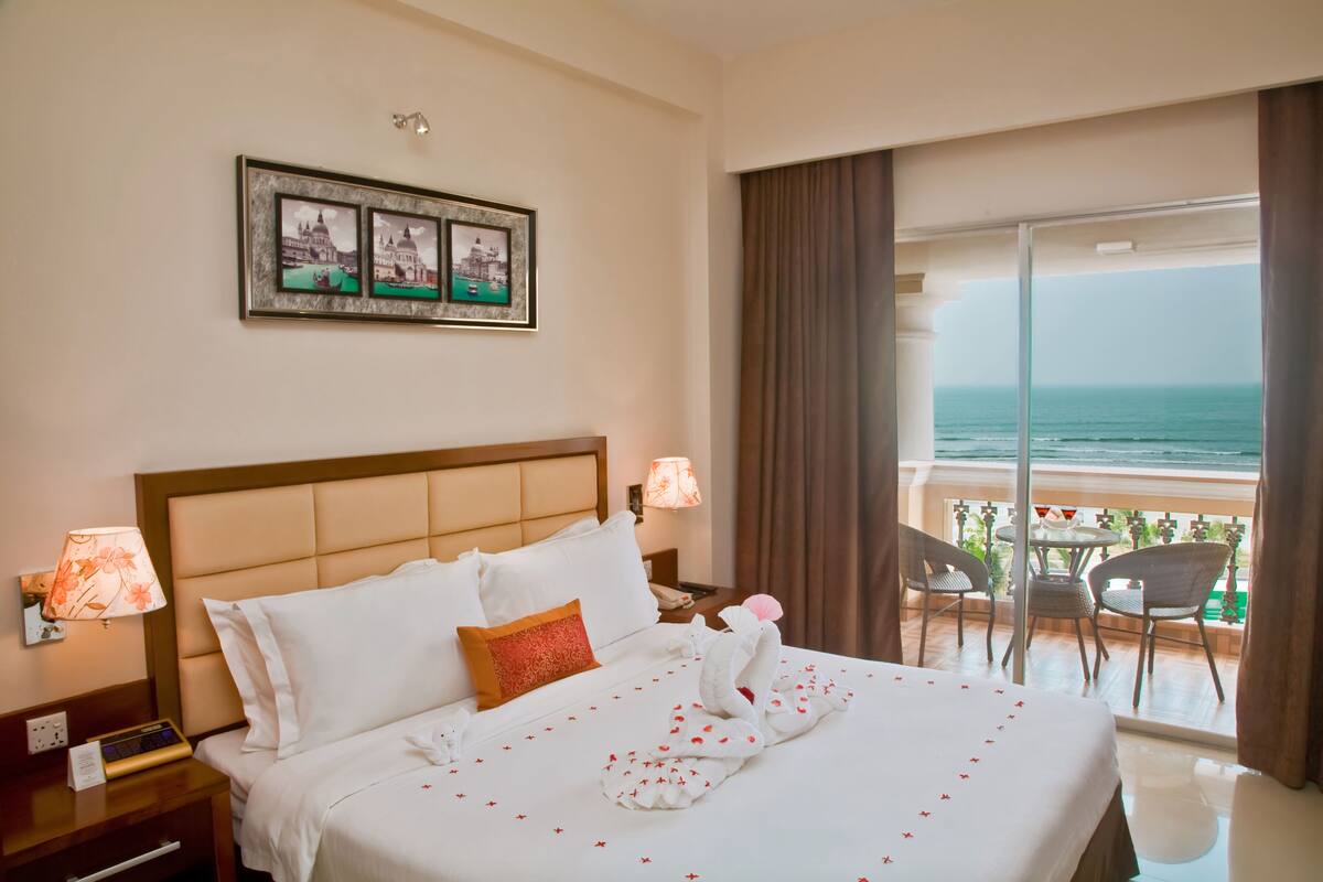 Une chambre d’hôtel avec un grand lit, une table de chevet avec une lampe, un tableau encadré au-dessus du lit et un balcon avec vue sur l’océan.