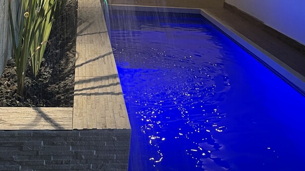 Une piscine dont le bord est illuminé de lumière bleue, entourée d’un mur blanc et d’une plante.