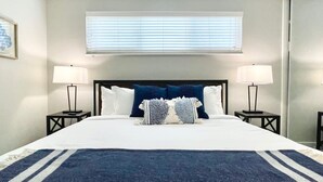 Una cama bien hecha con un edredón azul oscuro, dos lámparas de noche y una ventana con persianas.