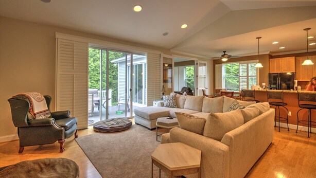 A spacious living room with a large sectional sofa, a leather armchair, and a dog bed. There is a sliding glass door leading to a patio with outdoor furniture. The kitchen area is visible with a bar and a refrigerator. - 4 BR, 5 BA Home, Pet Friendly, EV Charger, Community Pool, Tennis, Free Activities and Chair Lift! (Selbyville)