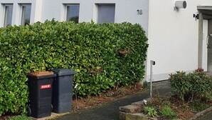 Il s’agit d’un immeuble résidentiel de deux étages, avec un toit gris, des murs blancs et le chiffre « 18 » peint sur le côté. Devant l’immeuble, on trouve une poubelle noire portant l’inscription « SULO ».
