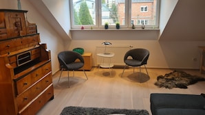 Une chambre de style loft moderne avec un plafond en pente, une commode en bois, deux chaises, une petite table et une vue sur les bâtiments à travers de grandes fenêtres.