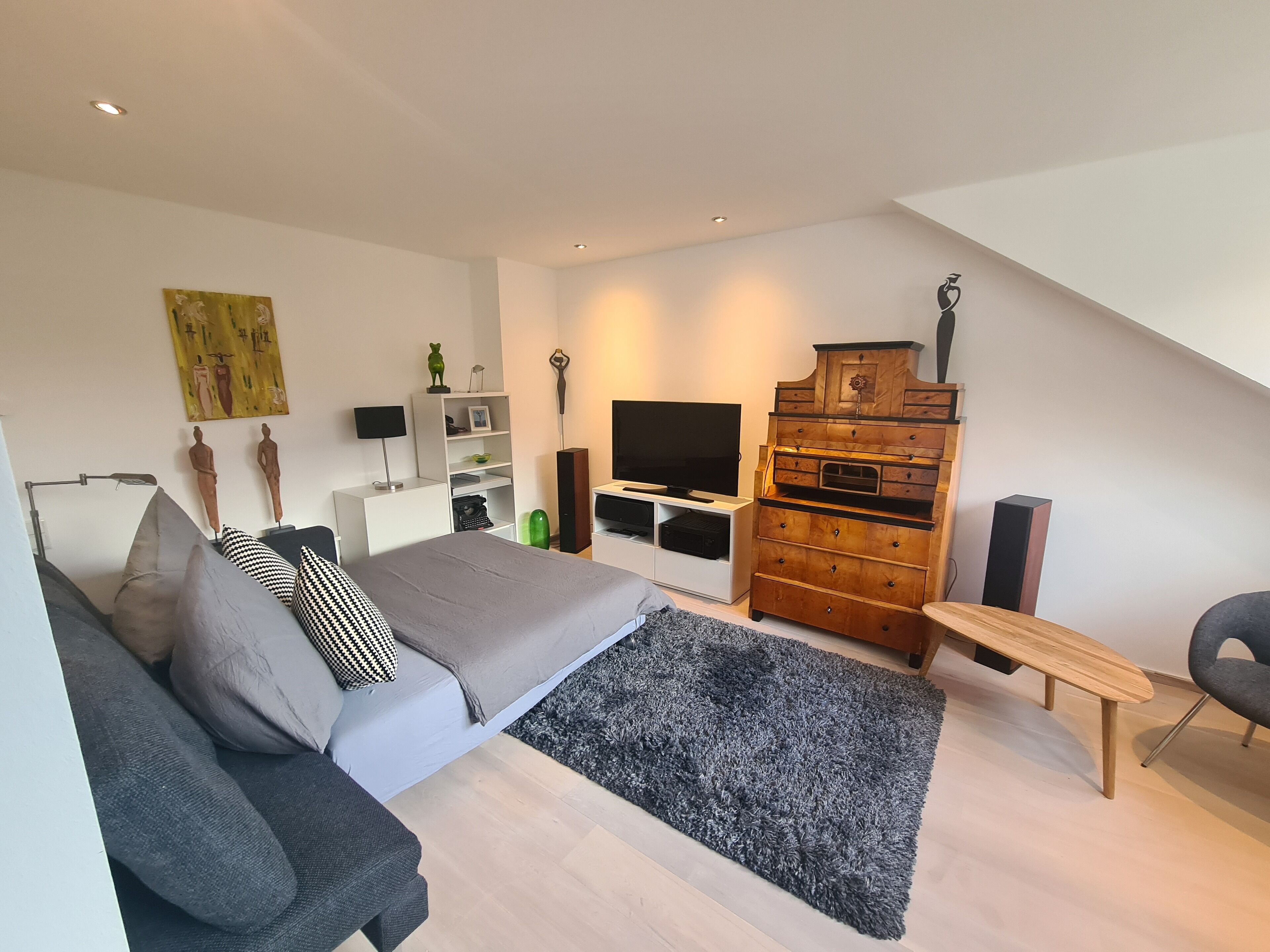 Ein modernes Schlafzimmer mit einem Bett, einem grauen Sofa, einer Holzkommode, einem Fernseher und einem kleinen Tisch.