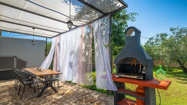 An outdoor patio with a stone floor, a wooden table with chairs, a grill, and a pergola with white curtains. - Holiday House Chill out Loborika (Marcana)