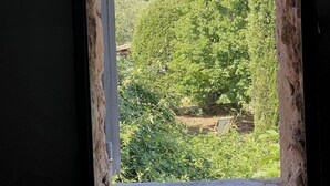 Une fenêtre donnant sur un paysage verdoyant et luxuriant, avec des arbres et, au loin, une colline surmontée d’un bâtiment au toit rouge.
