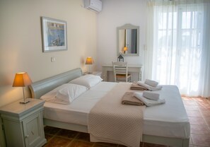 A neatly made bed with pillows and a blanket, a bedside table with a lamp, a desk with a chair, a mirror, and a window with curtains.
