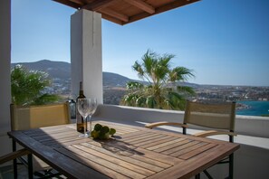 Une table en bois, sur laquelle se trouvent une bouteille et un verre, est placée sur un balcon donnant sur un paysage côtier, avec des palmiers et des montagnes en toile de fond.