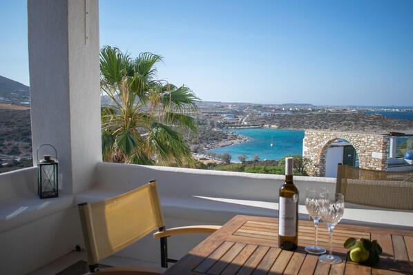 Un balcon avec une table, une bouteille de vin et des verres, donnant sur un paysage côtier avec un bâtiment en pierre et des palmiers.