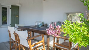 Een eethoek met een houten tafel en stoelen, een raam met groene luiken en een potplant met roze bloemen.