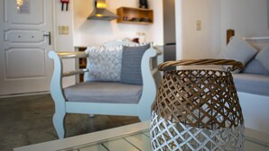 A modern living room with a white sofa, a wicker side table, and a kitchen area in the background. - Villa 78 m2 in Agia Irini, 350 Meter to the Beach for 4 Guests With Pool Access! (Paros)