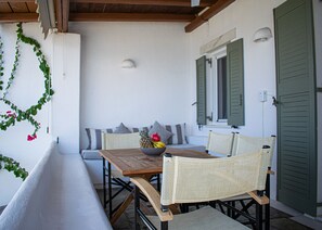 A covered outdoor seating area with a wooden table and wicker chairs, a bowl of fruit, and a bench. - Villa 78 m2 in Agia Irini, 350 Meter to the Beach for 4 Guests With Pool Access! (Paros)