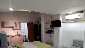 A bedroom with a bed, a small desk, a chair, a TV, and a kitchenette in the background.