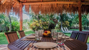Una mesa de madera preparada para una comida, bajo un techo de paja, rodeada de vegetación exuberante.