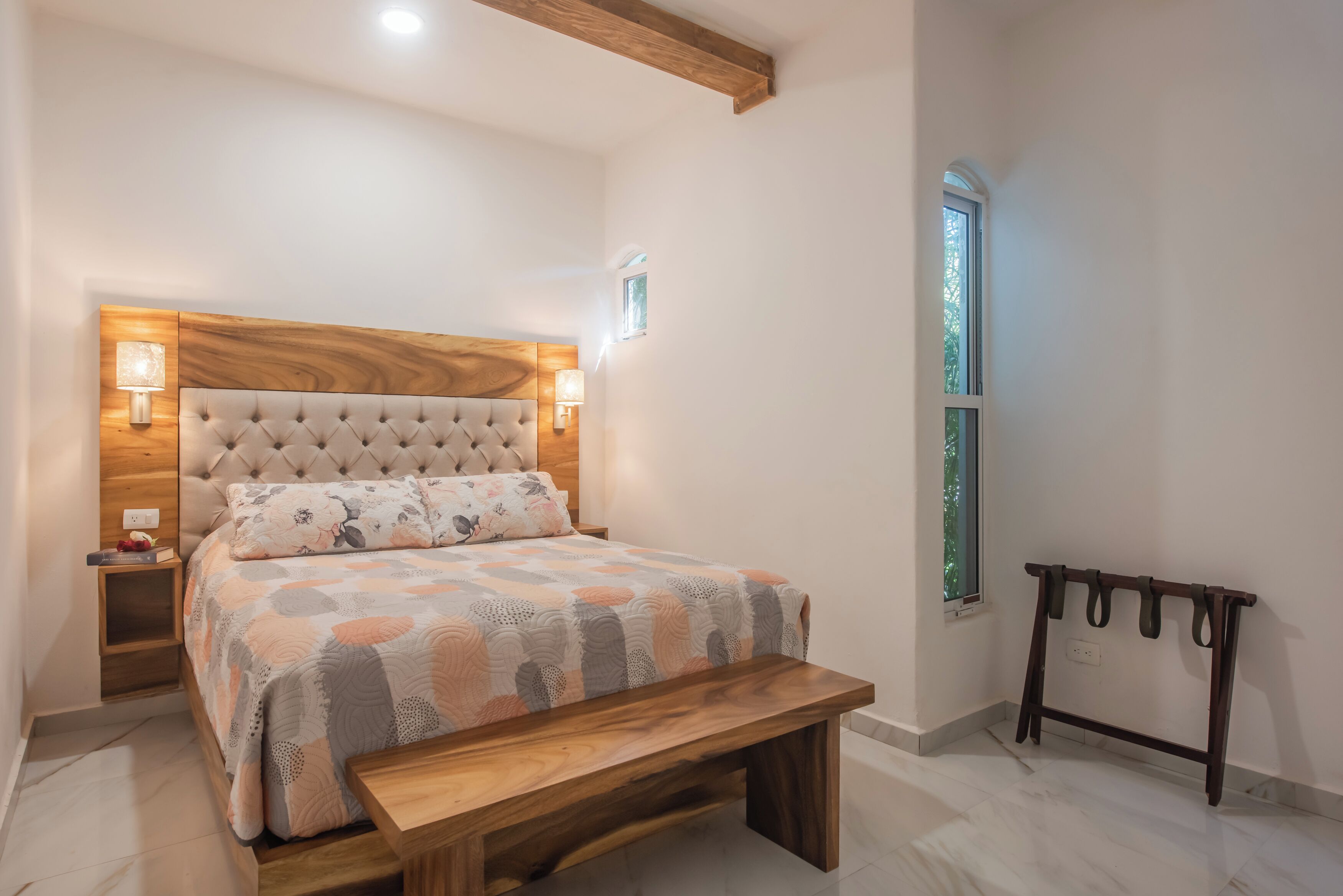 A bedroom with a tufted headboard, a wooden bench, a bedside table, and a wall-mounted light fixture.