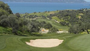 En golfbana med en sandbunker, träd och utsikt över havet och bergen i bakgrunden.
