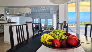 A modern kitchen with a central island, a dining area with a fruit bowl, and a view of the ocean. - Villa Pastorale-360 Degree View-boule Game (Paros)