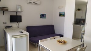 A compact kitchenette with a refrigerator, a microwave, and a small sink. There is a purple sofa and a white table with a bowl of food. A television is mounted on the wall, and there are two framed pictures on the wall. - lu Ientu House in Otranto, Baia dei Turchi Area No001 (Otranto)