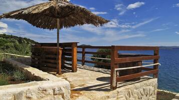A wooden deck with a thatched umbrella, stone walls, and a view of the sea.