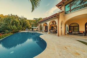 Ein Pool mit gewölbter Kante, umgeben von einer überdachten Terrasse mit Sitzgelegenheiten und Blick auf eine Hügellandschaft.