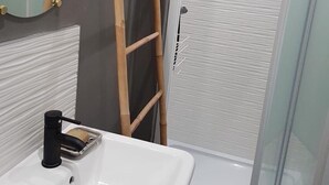 Ein Badezimmer mit Waschbecken in Weiß, einem Spiegel, einer Dusche mit Glastür und einer Holzleiter, die an der Wand lehnt.