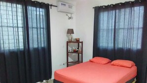 A bedroom with a bed, black curtains, a small table with a lamp, and a wall-mounted air conditioner.