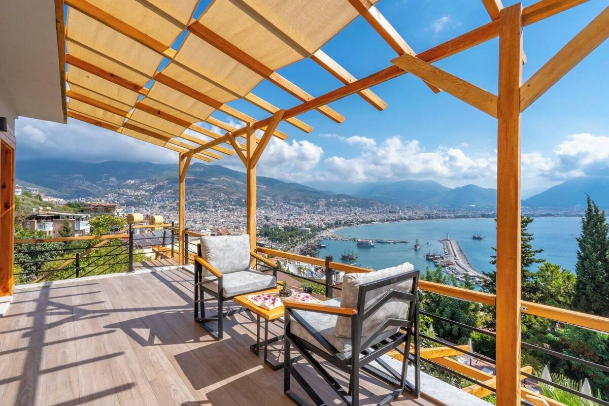Une terrasse meublée de mobilier en bois, une vue sur une ville côtière et un ciel dégagé.