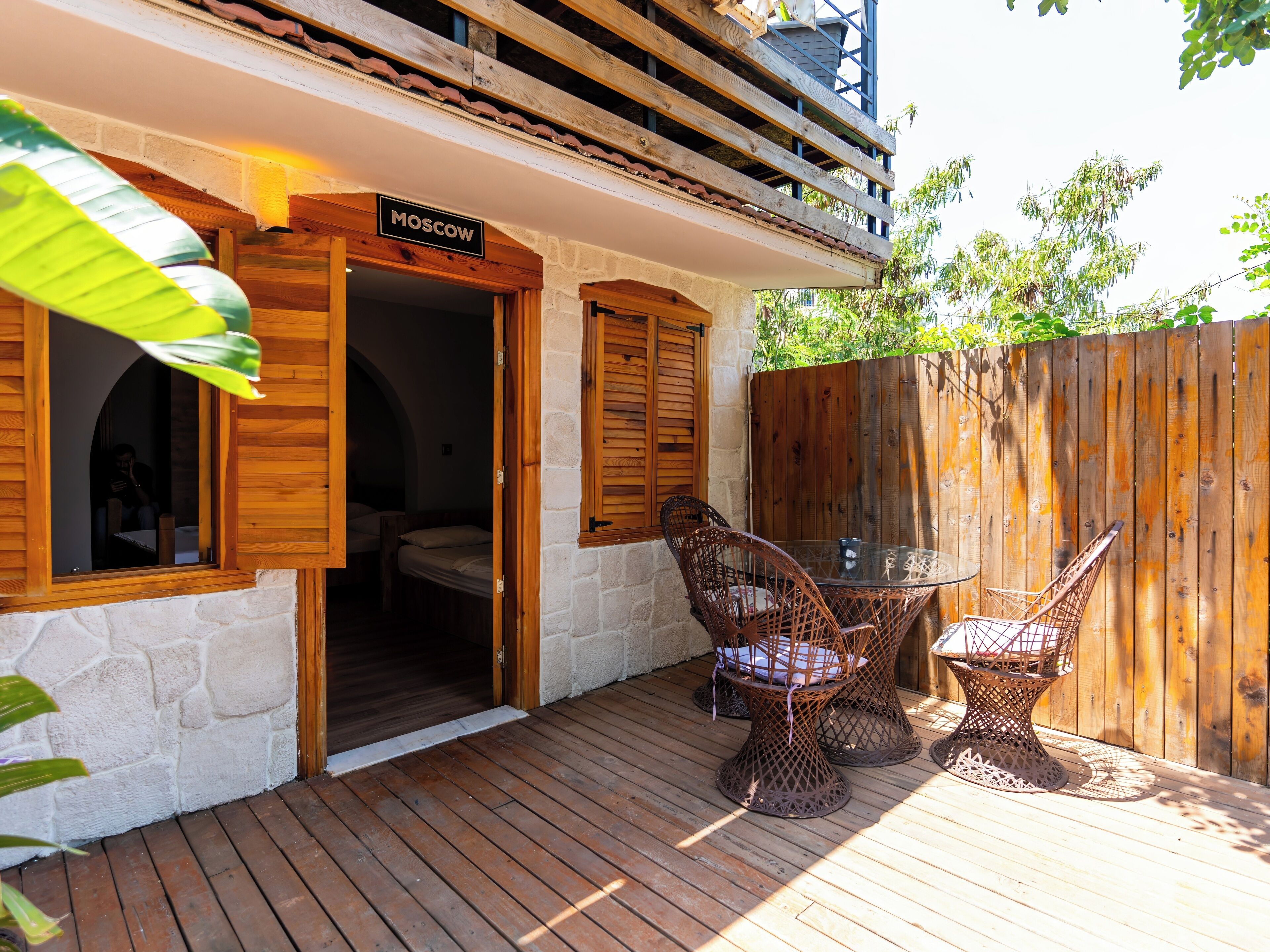 Une terrasse en bois avec des chaises et une table en osier, un panneau indiquant « Moscou », et un mur en pierre.