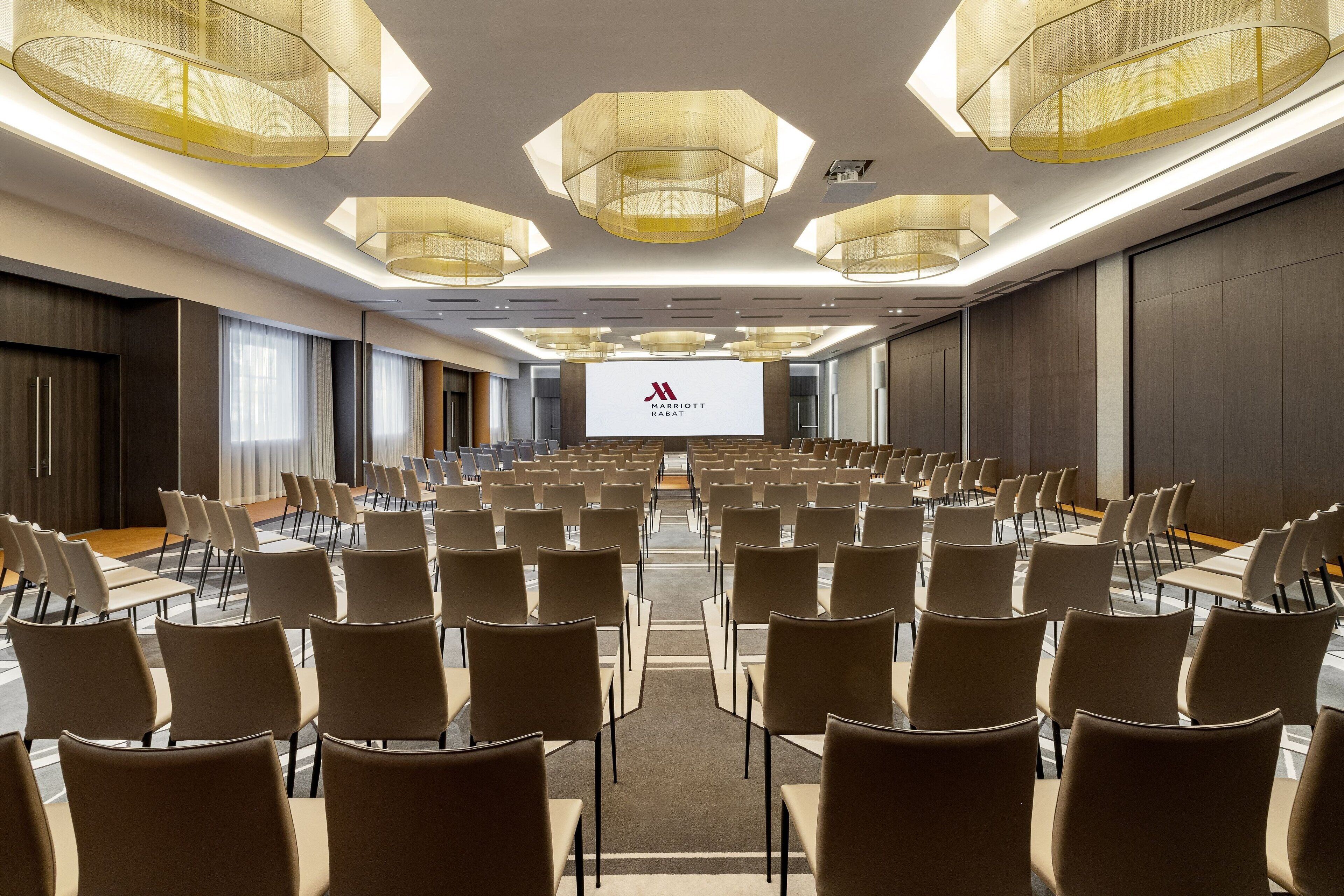 A conference room with rows of chairs, a large screen at the front, and modern chandeliers.