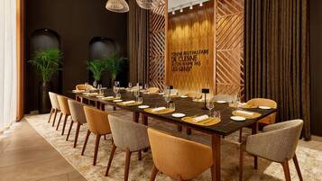 A dining area with a long table set for a meal, surrounded by chairs, with a decorative wall and plants in the background.