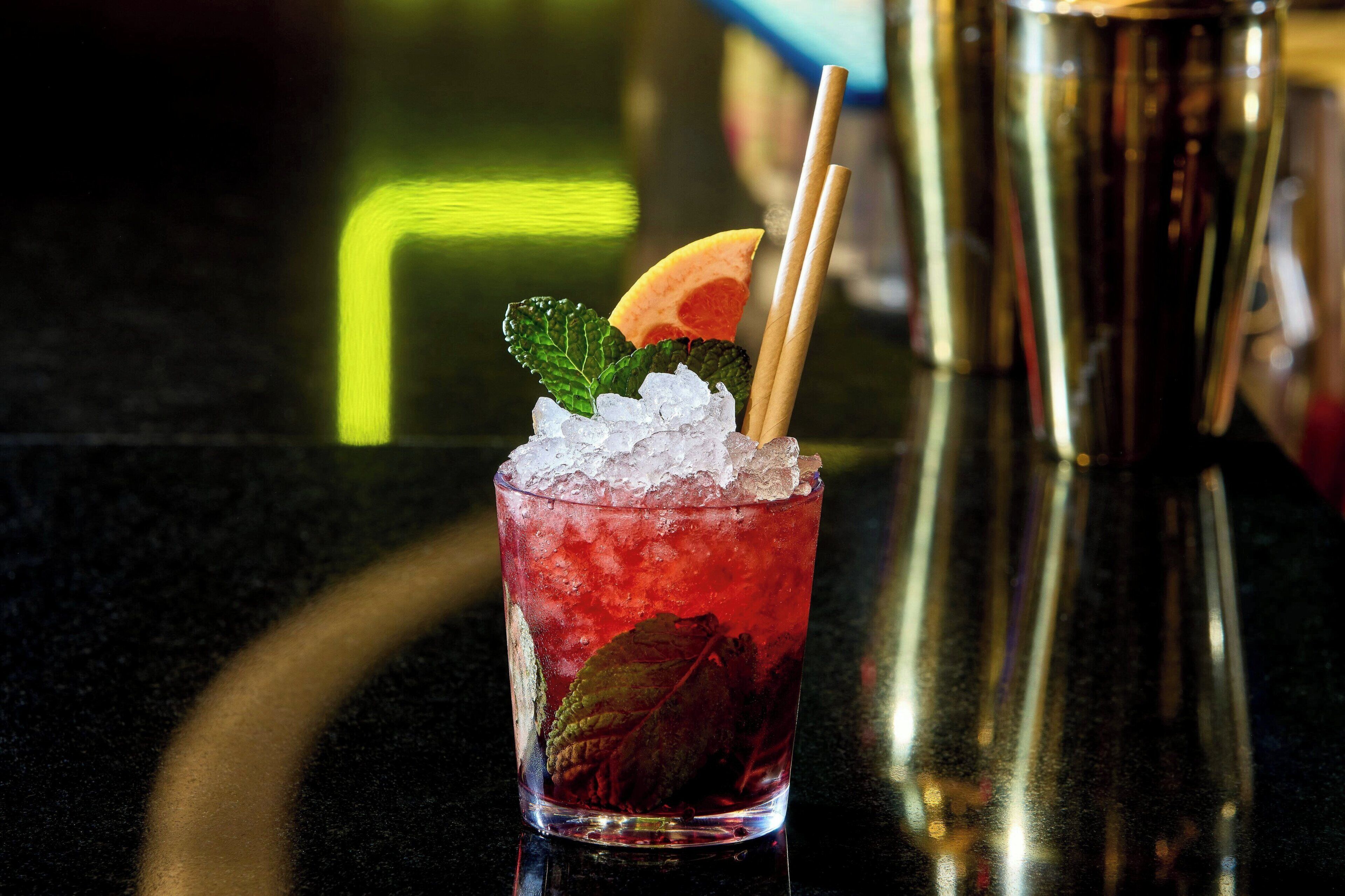 Een cocktail met ijs, een stuk fruit en een muntblaadje in een glas, met een cocktailshaker op de achtergrond.