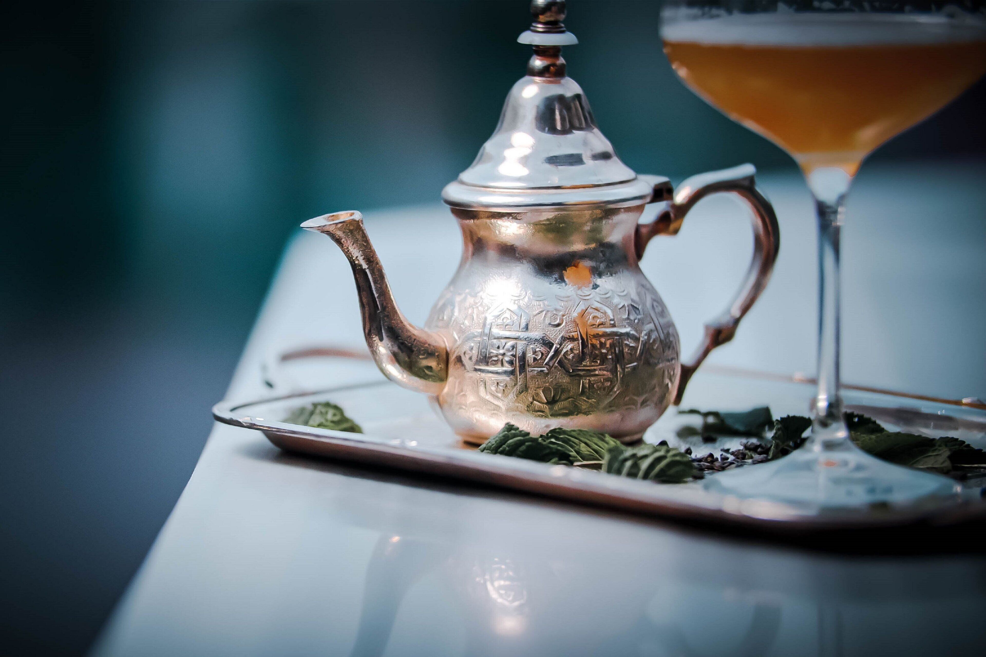 A silver teapot with intricate designs on a tray with tea leaves and a glass of amber-colored beverage.