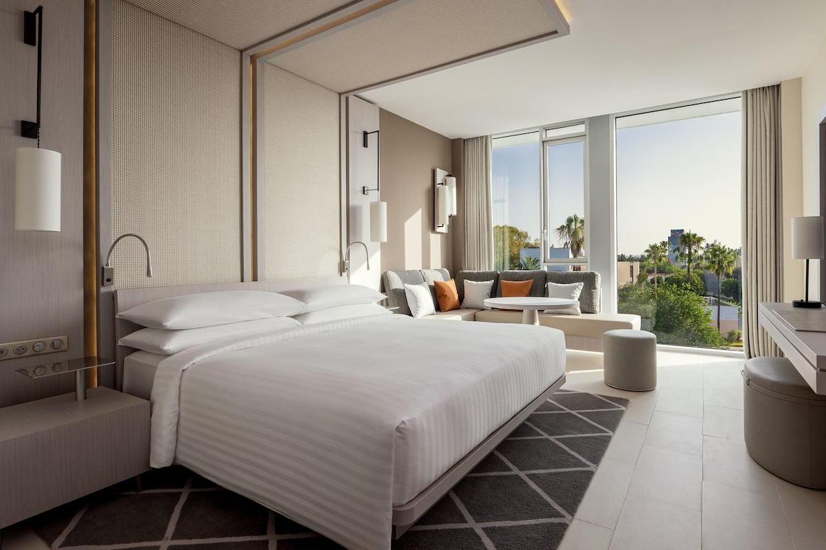 A modern hotel room with a large bed, a seating area with a sofa and armchairs, a small table, and a view of the city through large windows.