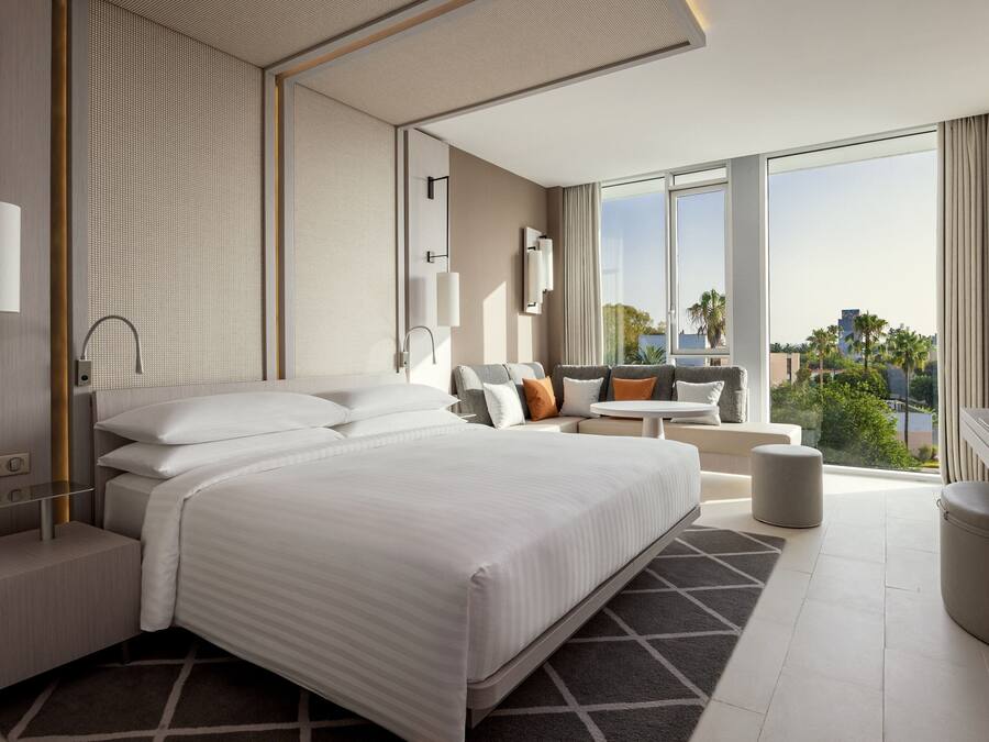 Une chambre d’hôtel moderne dotée d’un grand lit, d’un coin salon avec un canapé et des fauteuils, d’une petite table et offrant une vue sur la ville grâce à de grandes fenêtres.