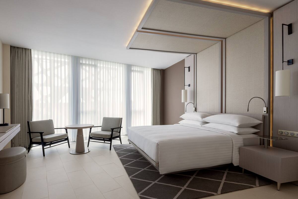 A modern hotel room with a large bed, a desk, two chairs, a small table, and a large window with sheer curtains.