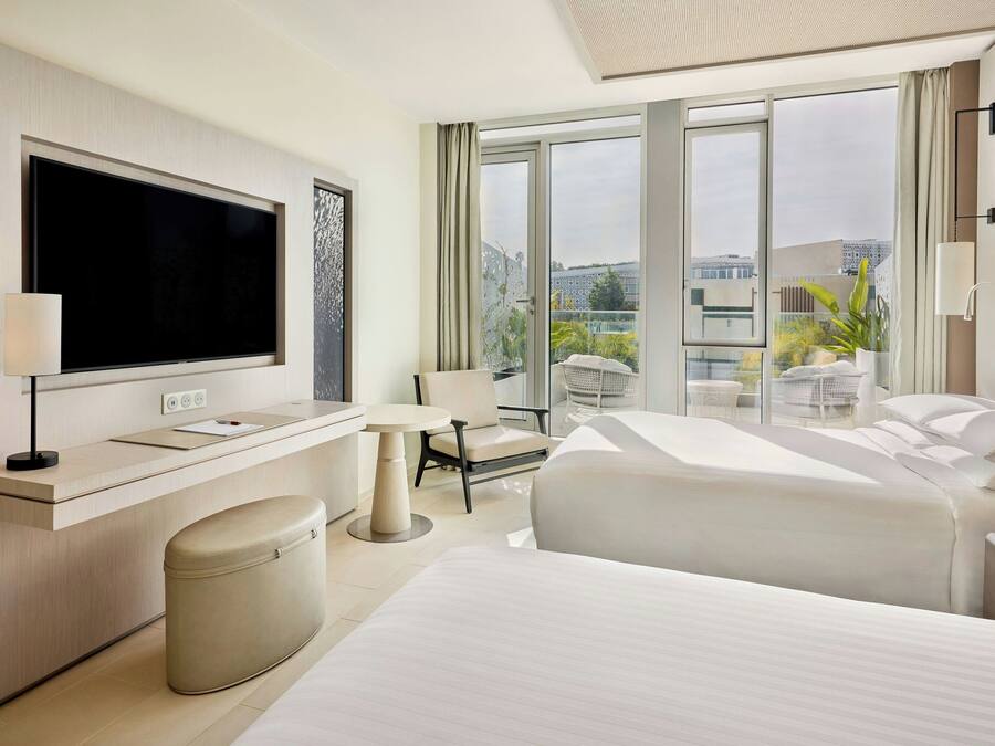 A modern hotel room with a large flat-screen TV, a desk with a lamp, a comfortable chair, a bed, and a view of the city through large windows.