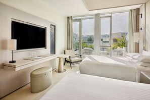 A modern hotel room with a large flat-screen TV, a desk with a lamp, a comfortable chair, a bed, and a view of the city through large windows.