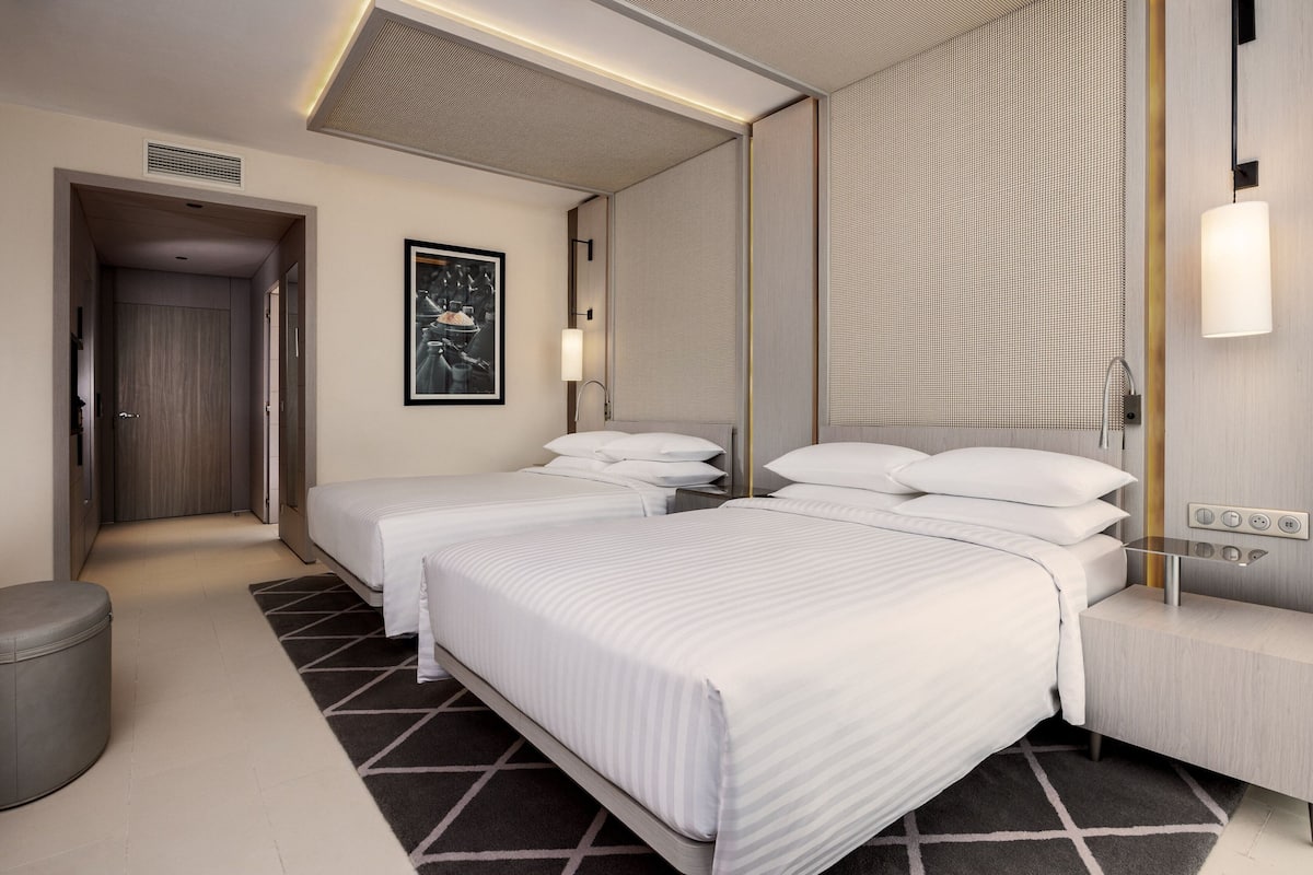 A modern hotel room with two beds, a large mirror, a bedside table with a lamp, and a small seating area.