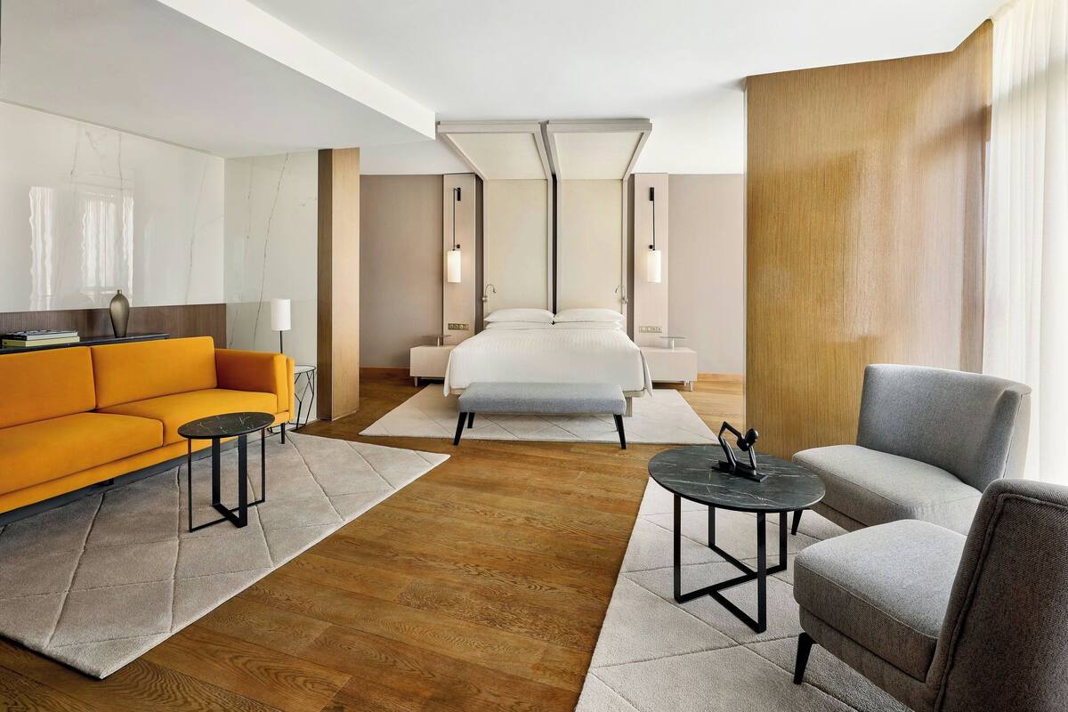 A modern hotel room with a bed, a sofa, a chair, a small table, and a lamp.