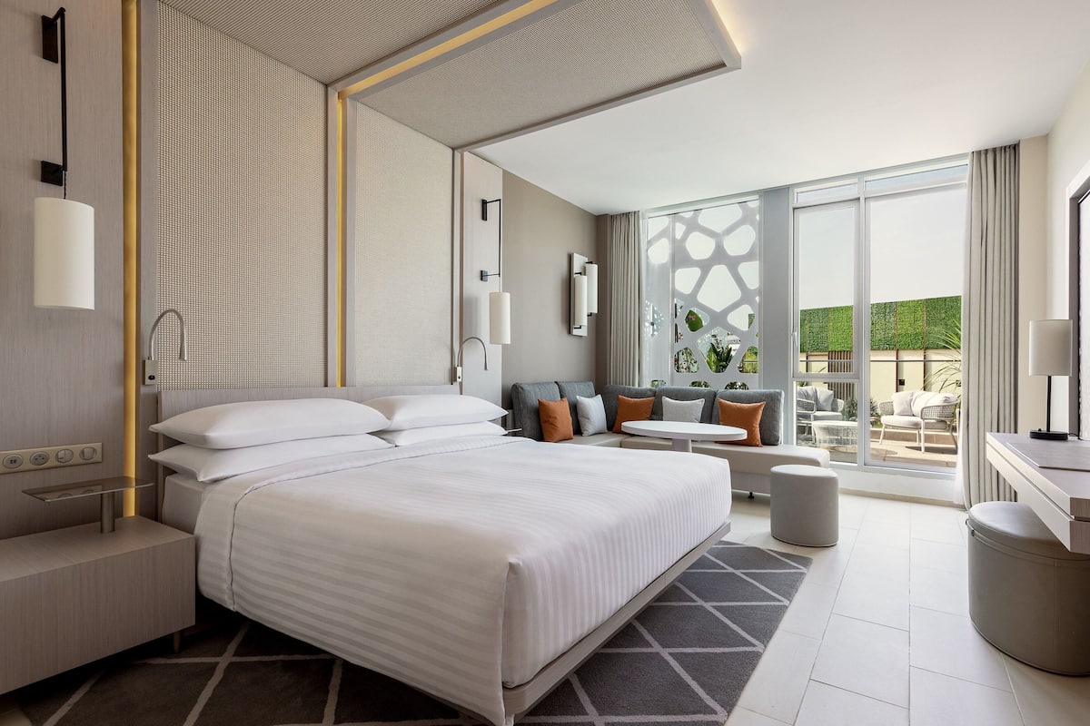 A modern hotel room with a large bed, a desk, and a seating area with a view of the outdoors.
