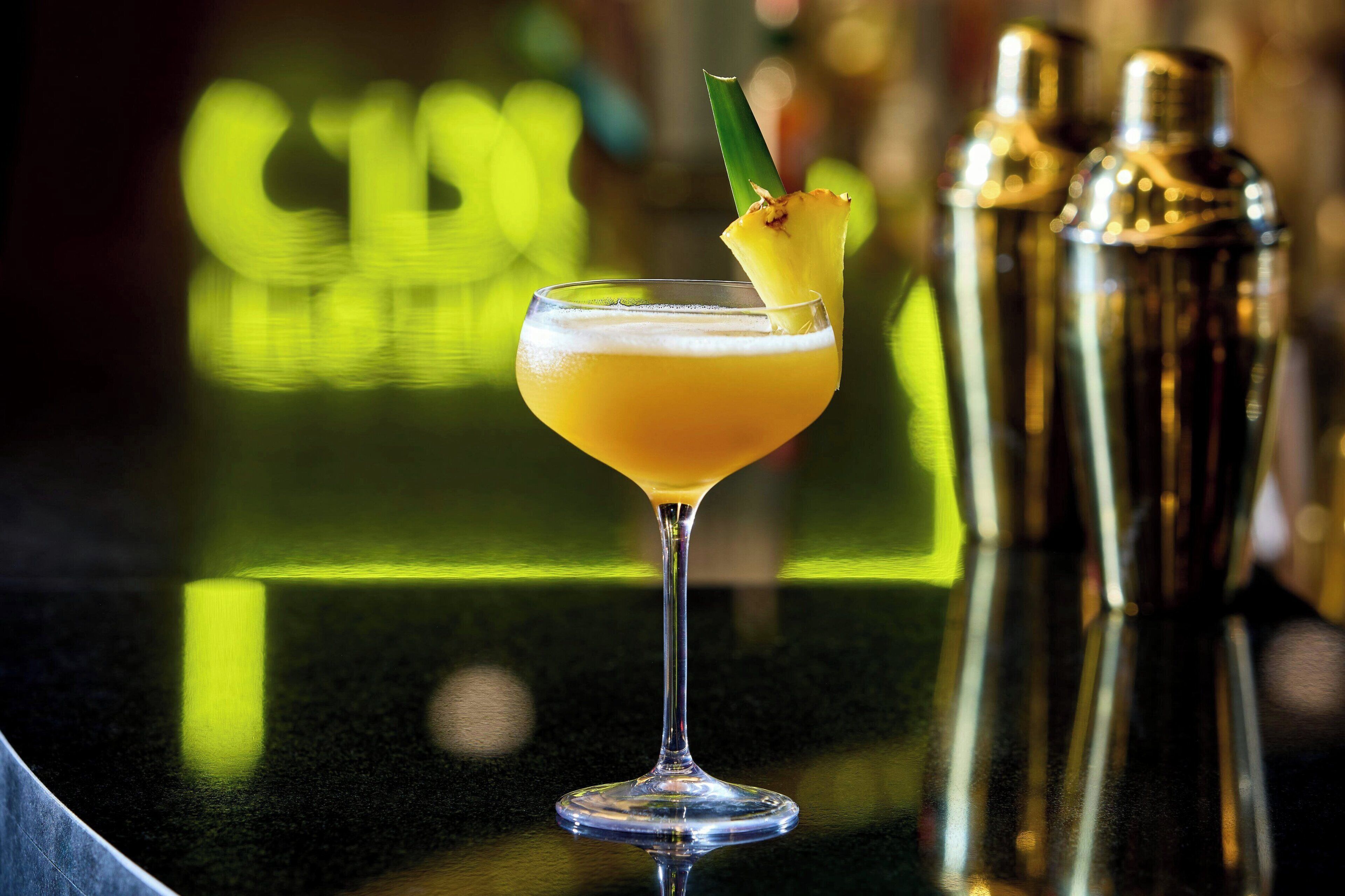 A cocktail in a stemmed glass with a pineapple garnish, placed on a reflective bar surface.