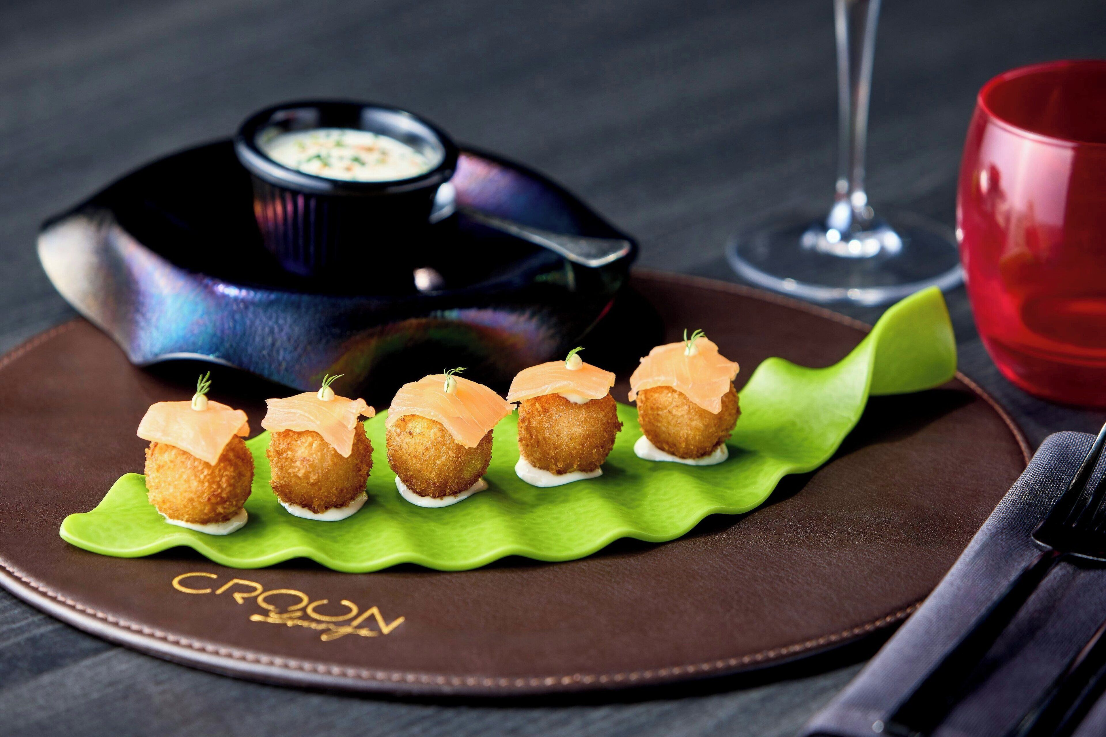 A plate with croquettes and a dipping sauce, a glass of red wine, and a red cup on a table.