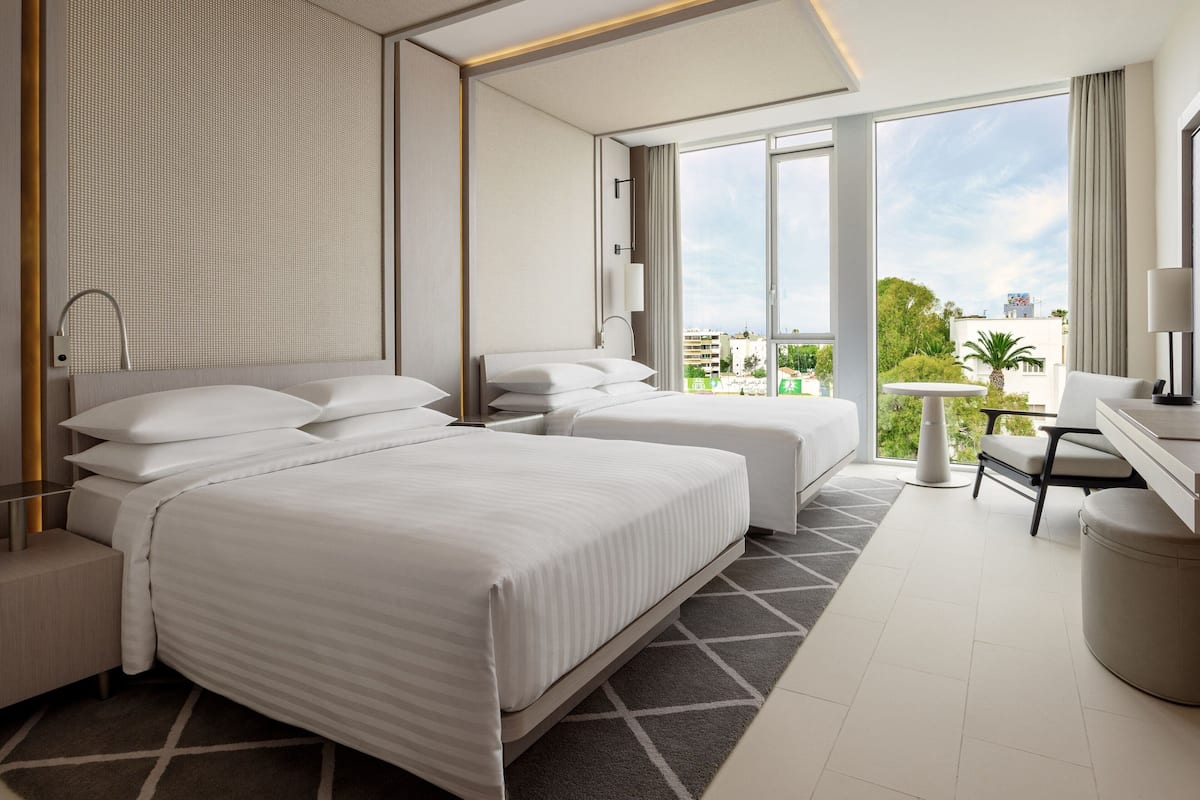 A modern hotel room with two beds, a desk, and a view of the city.
