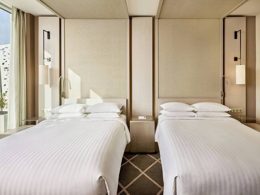 A hotel room with two beds, a large window with curtains, a small table, and a wall-mounted light fixture.