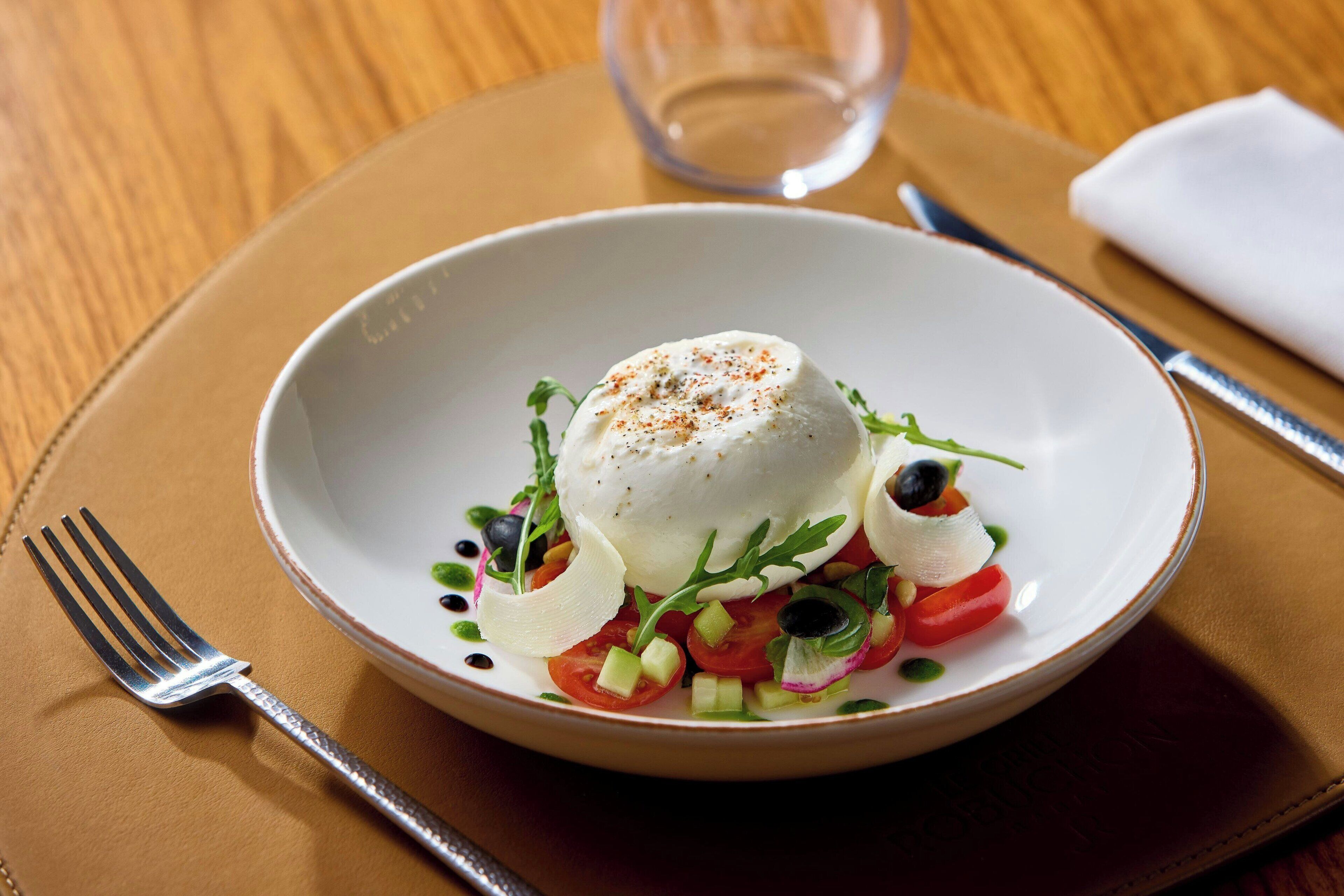 A white plate with a white food item, garnished with green leaves and black olives, on a brown tablecloth.