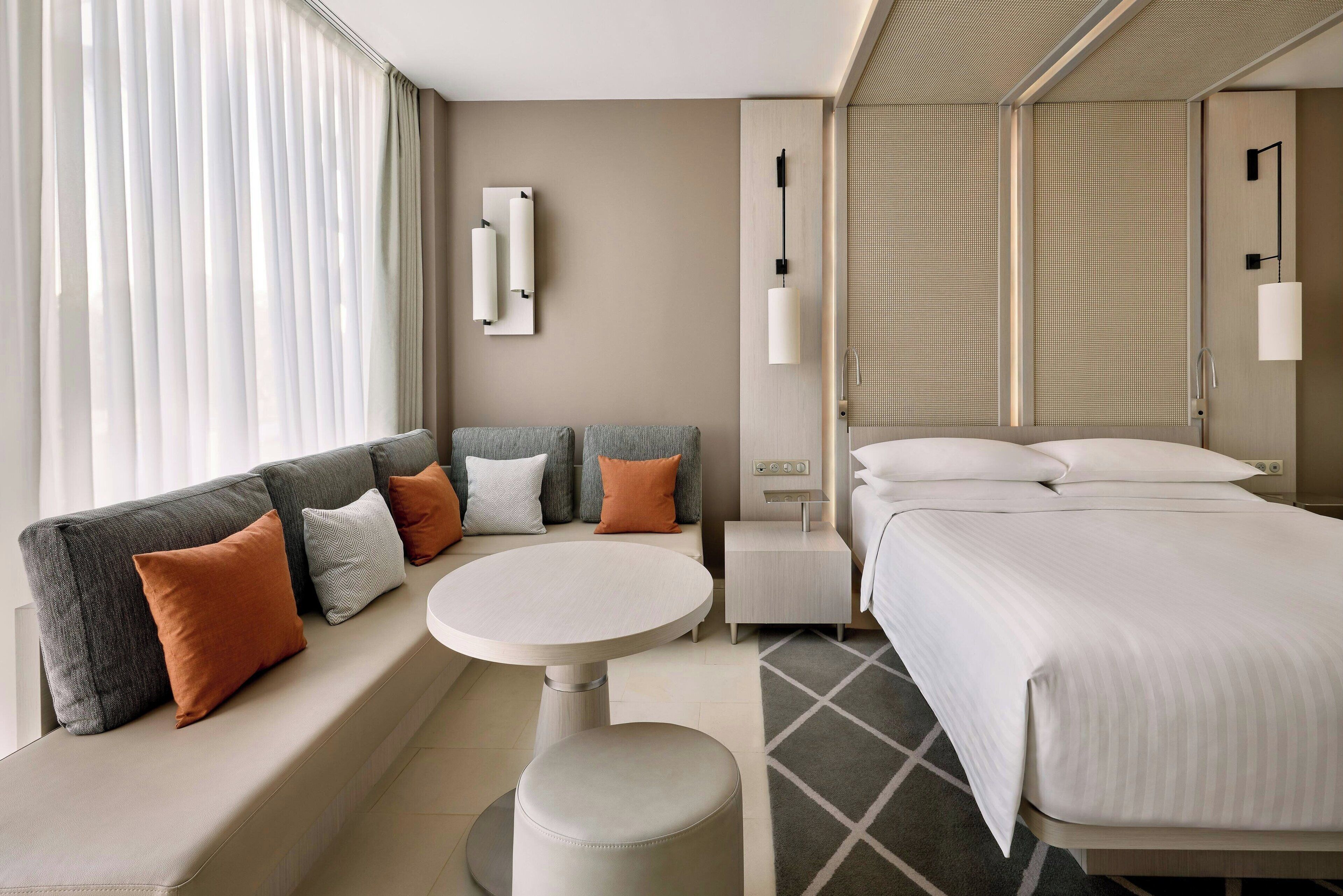 A modern hotel room with a sofa, a round table, a bed, and a small bedside table.
