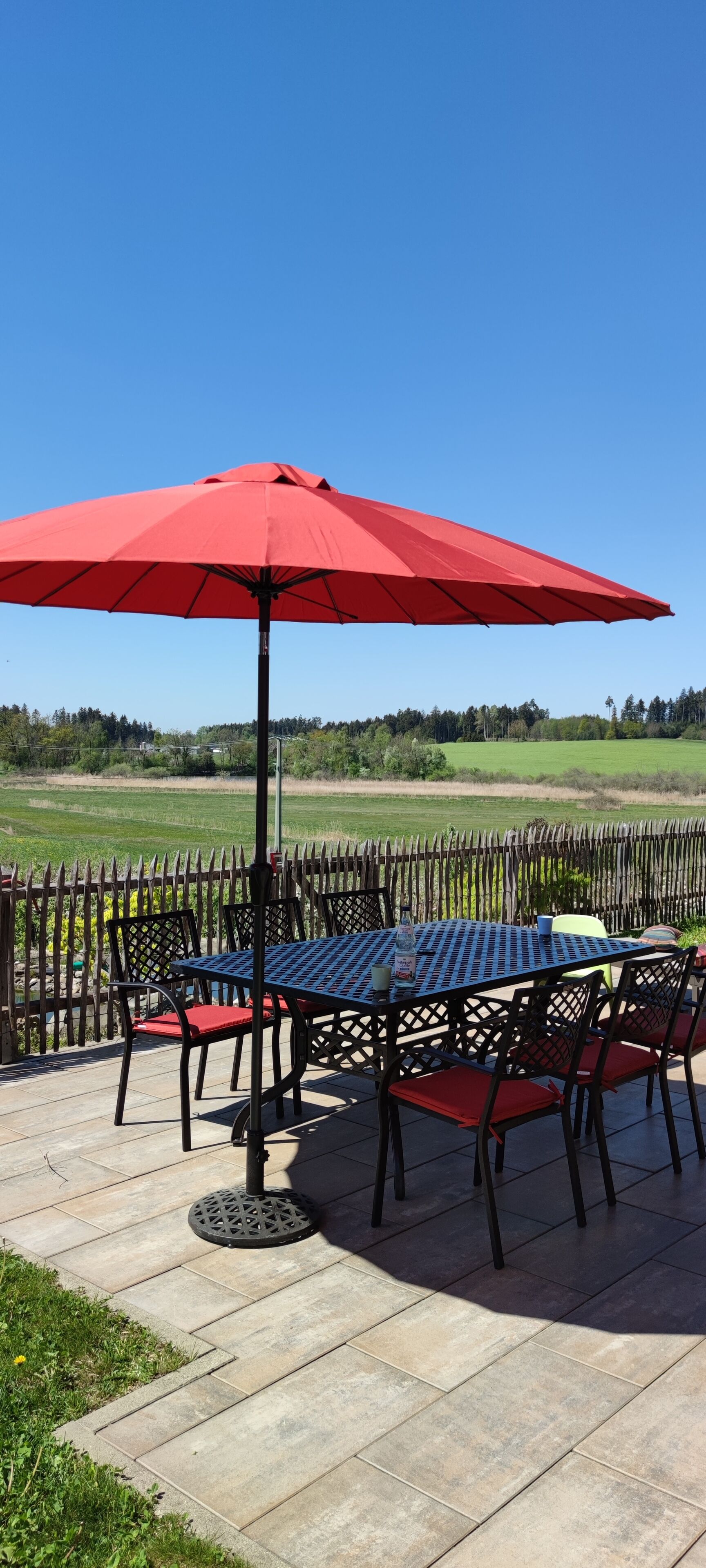 Een terras buiten met een tafel, stoelen, een rode parasol en uitzicht op een groen veld.