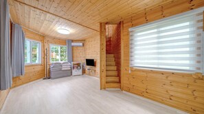 A room with wooden walls and ceiling, a large window with blinds, a television on a stand, and a stack of pillows. - Paju Gamaksan Sanchon Village Pension (Paju)